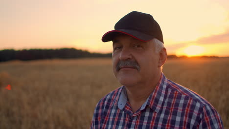 Portrait-of-a-happy-Senior-adult-farmer-in-a-cap-in-a-field-of-grain-looking-at-the-sunset.-Wheat-field-of-cereals-at-sunset.-Slow-motion